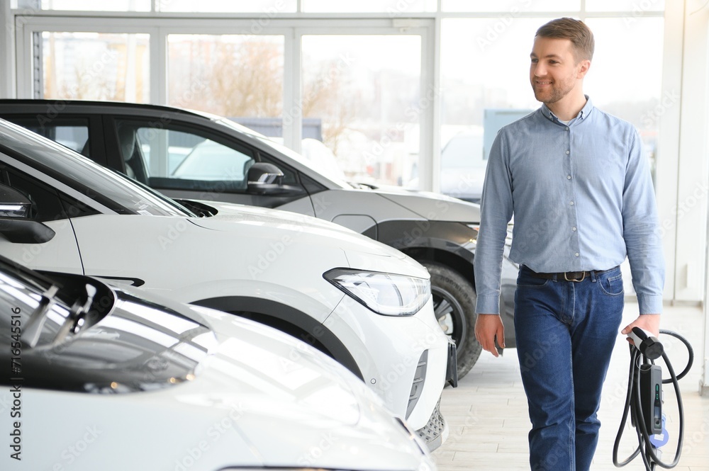 Handsome business man holding charging cable for electric car. Caucasian male stands near electric auto in dealership