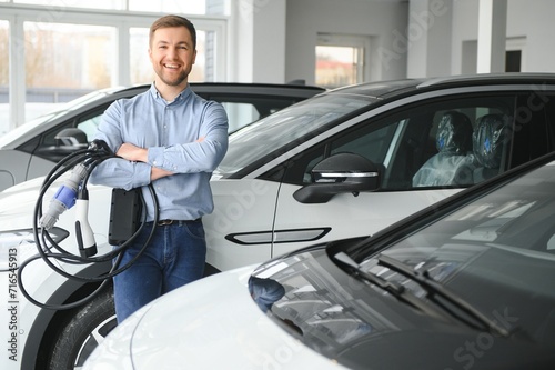 Concept of buying electric vehicle. Handsome business man stands near electric car at dealership