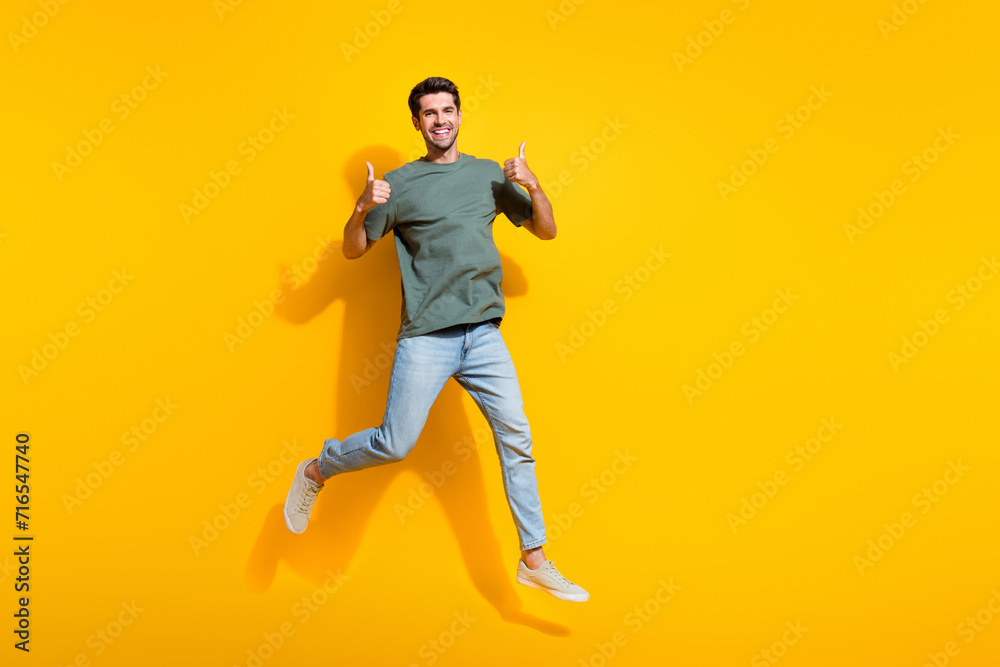 Full length photo of confident cheerful man dressed khaki t-shirt showing thumbs up jumping empty space isolated yellow color background