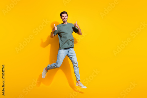 Full length photo of confident cheerful man dressed khaki t-shirt showing thumbs up jumping empty space isolated yellow color background photo