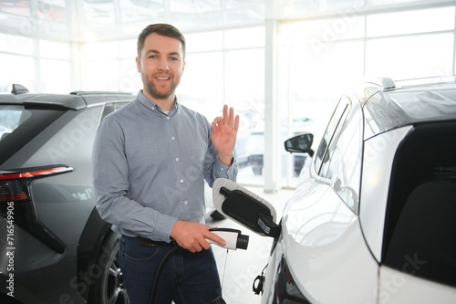 Concept of buying electric vehicle. Handsome business man stands near electric car at dealership © Serhii
