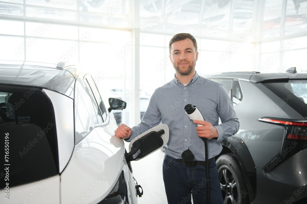 Young man buying first electric car in the showroom. Eco car sale concept Young man buying first electric car in the showroom. Eco car sale concept