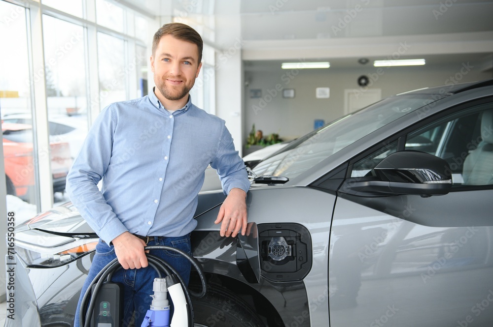 Concept of buying electric vehicle. Handsome business man stands near electric car at dealership