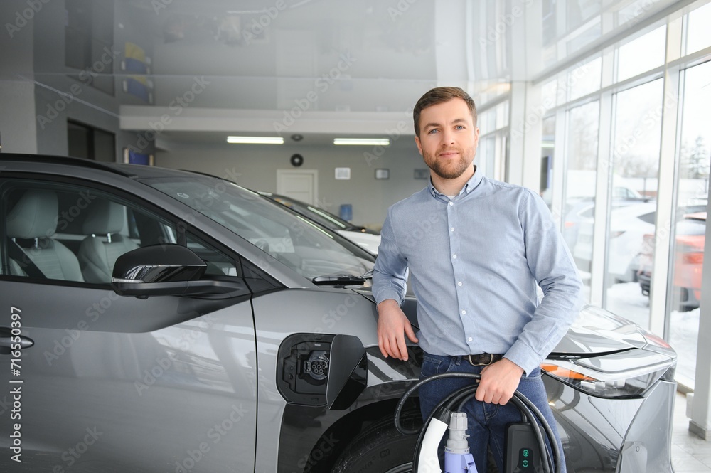 Young man, selling electric cars in the showroom. Concept of buying eco-friendly car for family