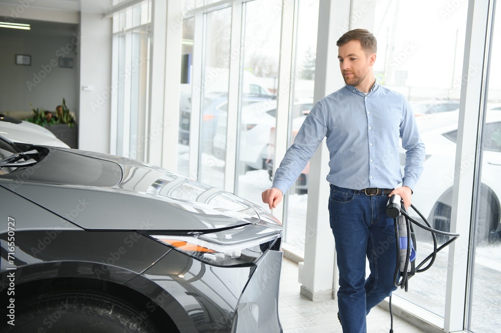 Obraz premium Handsome business man holding charging cable for electric car. Caucasian male stands near electric auto in dealership