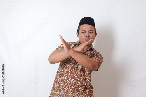 Portrait of excited Asian man wearing batik shirt and songkok  showing X sign of hands, refusing an invitation from someone, making prohibitive gesture. Isolated image on gray background