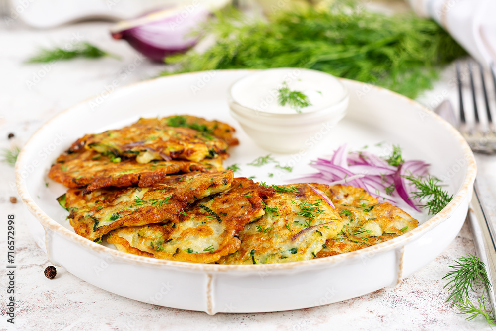 Zucchini fritters with red onions, garlic and herbs. Vegetarian zucchini pancakes and sour cream on white table. Zucchini fritters with red onions, garlic and herbs. Vegetarian zucchini pancakes and sour cream on white table.