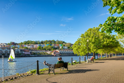 Bristol, UK - A beautiful spring evening at Bristol Docks, young couple sitting on a bench, colourful houses, fresh green trees, blue sky, sail boat on the water. photo