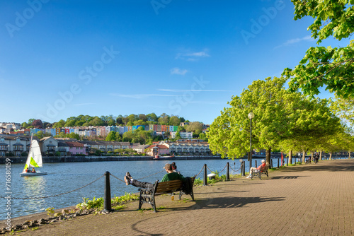 Bristol, UK - A beautiful spring evening at Bristol Docks, young couple sitting on a bench, colourful houses, fresh green trees, blue sky, sail boat on the water.
