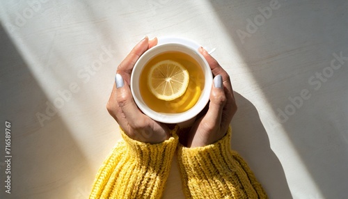 top view woman s hands holding a white cup of tea with a slice of lemon she is wearing a yellow cozy sweater the space is filled with warm light with soft shadows and copy space  photo