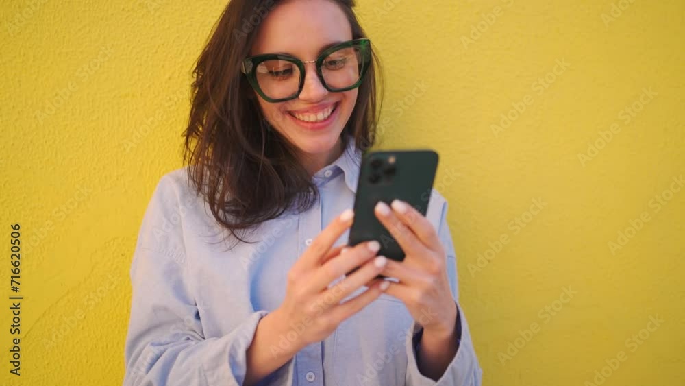 Smiling young woman with brown hair using smartphone against yellow wall. Cheerful female in eyeglasses reading messages on mobile phone. Freelancer in shirt looking at device screen with smile. Smiling young woman with brown hair using smartphone against yellow wall. Cheerful female in eyeglasses reading messages on mobile phone. Freelancer in shirt looking at device screen with smile.