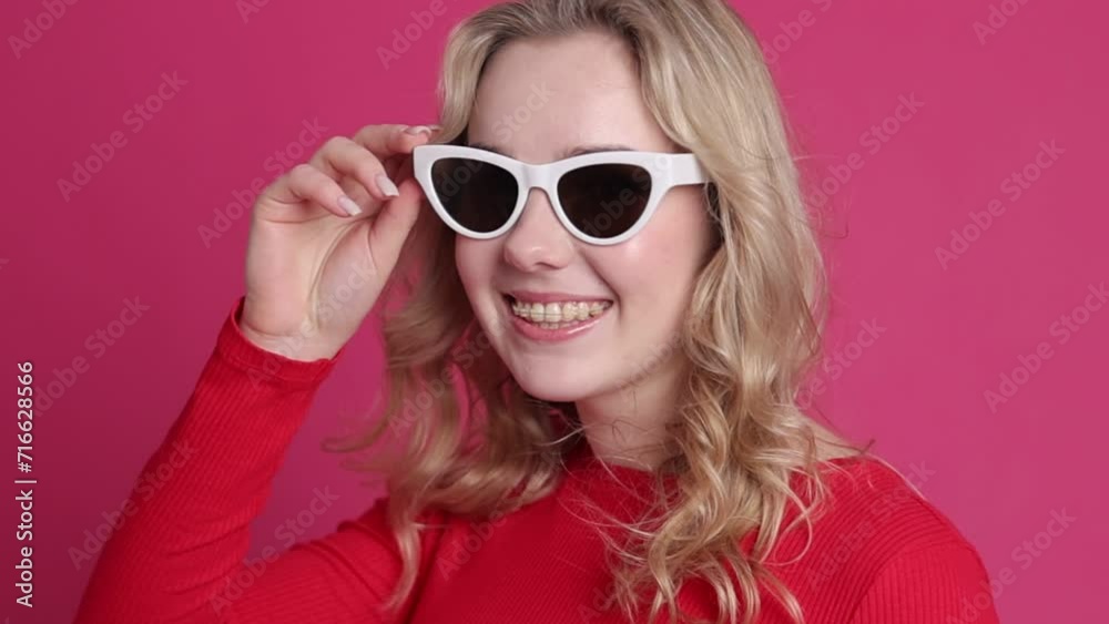 Smiling young caucasian blond woman with transparent ceramic braces on teeth isolated on pink backdrop. Girl takes off her stylish sunglasses and blinks at camera. Beauty, health and fashion concept. Smiling young caucasian blond woman with transparent ceramic braces on teeth isolated on pink backdrop. Girl takes off her stylish sunglasses and blinks at camera. Beauty, health and fashion concept.
