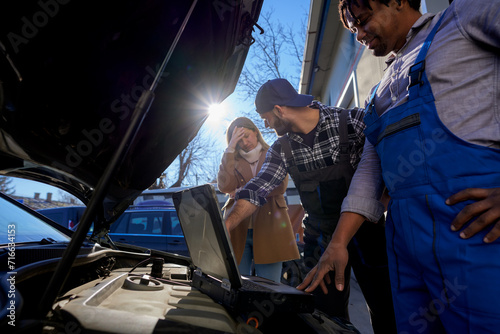 A worried woman, flanked by mechanic colleagues, scrutinizes a car using computer diagnostics. They engage in a detailed discussion about the issue at hand.