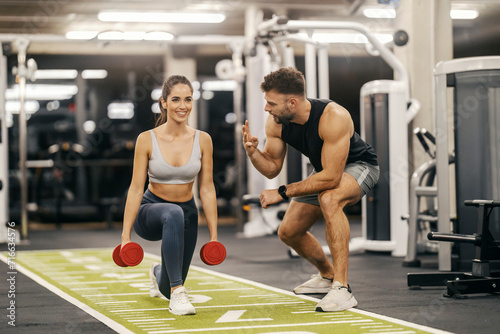 A fit sportswoman is doing lunges with dumbbells while her trainer is encouraging her.