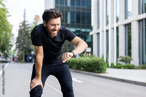 Fototapeta Naklejka Na Ścianę i Meble -  Young male athlete standing outside on elbow bent after jogging and marathon and looking disappointed and upset at smart watch on wrist, not happy with result