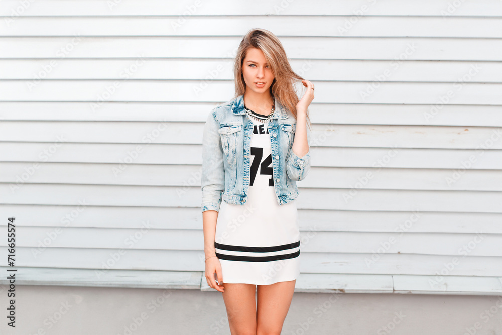 Beautiful young woman in fashion street denim clothes with a dress and a jeans jacket stands near a vintage wooden wall Beautiful young woman in fashion street denim clothes with a dress and a jeans jacket stands near a vintage wooden wall