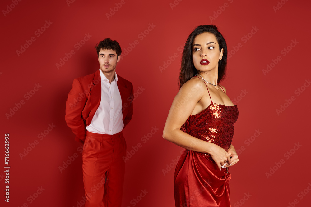 focus on gorgeous and young woman posing in sexy dress near blurred man in suit on red background focus on gorgeous and young woman posing in sexy dress near blurred man in suit on red background