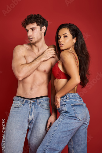 muscular and shirtless man in jeans standing with gorgeous brunette woman in bra on red backdrop