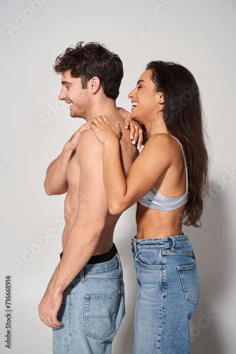 side view of happy young woman with brunette hair leaning on shirtless man on grey background