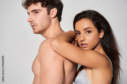 seductive young woman with brunette hair leaning on shirtless man on grey background, intimacy