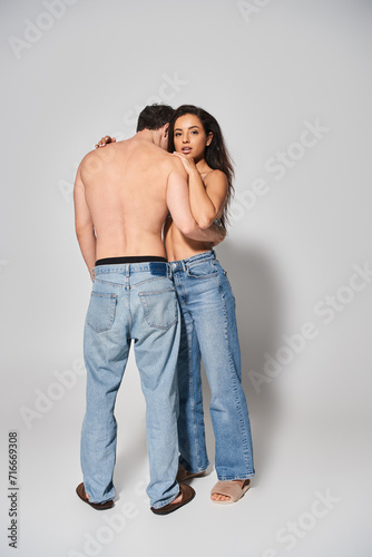 sexy and shirtless man hugging young woman in denim blue jeans on grey background, togetherness