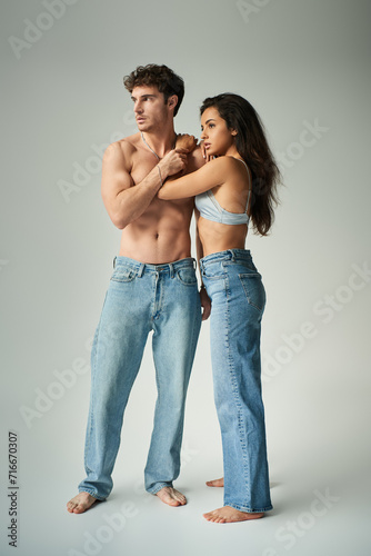 brunette young woman in satin bra and jeans embracing shirtless man on grey background, affection
