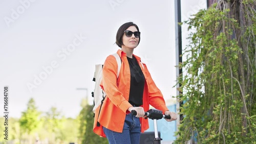 Smiling female in sunglasses riding e-scooter on road near residential buildings. Positive caucasian woman using contemporary ecological transport for moving throught city center.