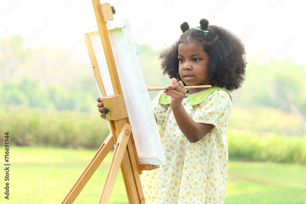 Cute smiling African girl with black curly hair painting on canvas at green garden outdoor. Kid drawing nature picture in green summer park. Happy little child having fun while spending time outdoors. - obrazy, fototapety, plakaty 