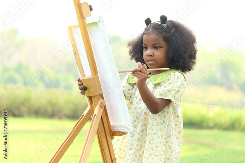 Bilde på lerret Cute smiling African girl with black curly hair painting on canvas at green garden outdoor