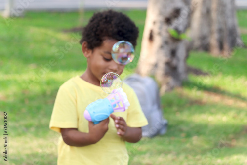 Soap bubble in blued green background, happy African boy making bubbles with blowing bubble gun toy in garden. Kid spends time outdoors in meadow. Child has fun with bubble in summer park. © Stella