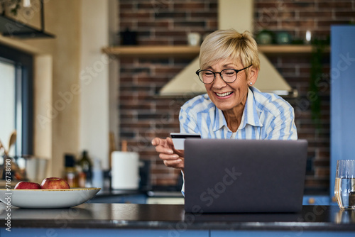 A smiling older woman, looking happy, shopping online, holding a credit card, using a laptop.