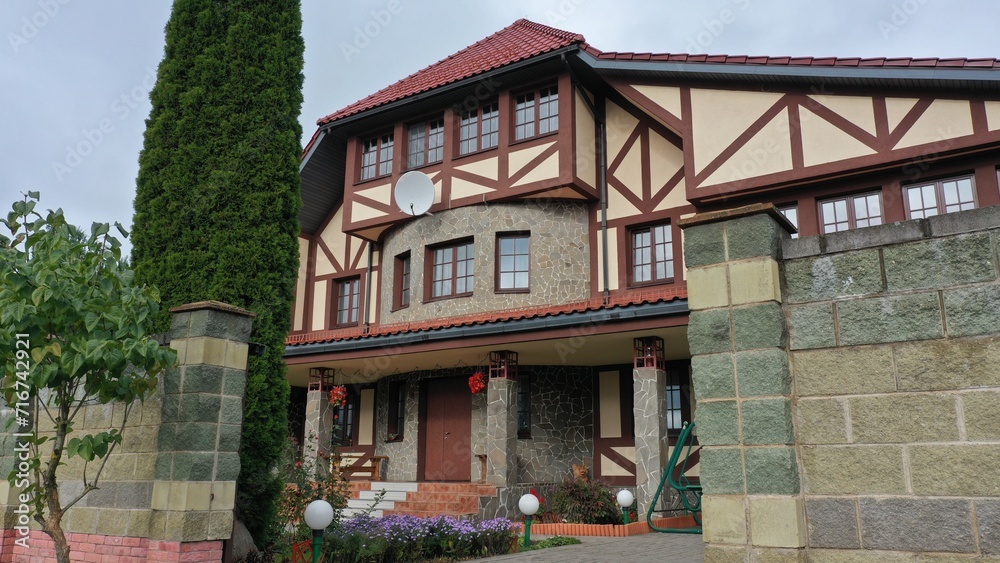 Naklejka premium Modern private three-story country house in English, European style with manicured green grass, stone wall, thuja fence and red tile roof. Pseudo-historic style of architecture.