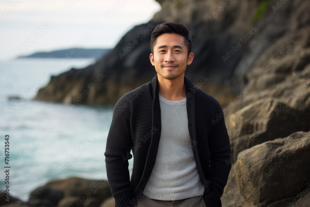 Portrait of a satisfied asian man in his 30s wearing a chic cardigan against a rocky shoreline background. AI Generation