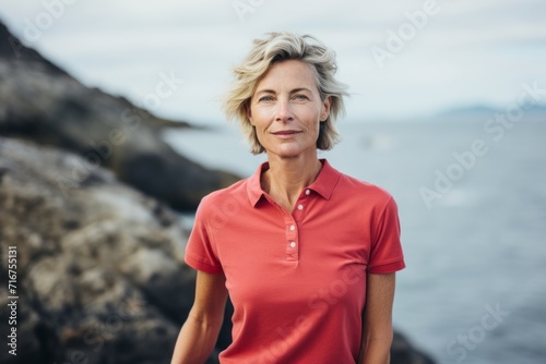 Portrait of a content woman in her 60s wearing a sporty polo shirt against a rocky shoreline background. AI Generation