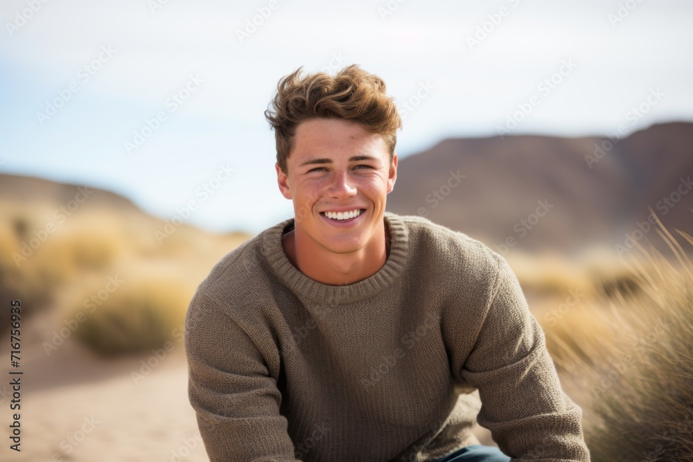 Portrait of a grinning man in his 20s wearing a cozy sweater against a serene dune landscape background. AI Generation