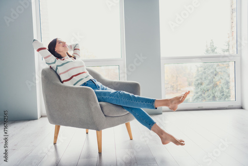 Full body profile photo of lovely satisfied glad lady sit barefoot chair hands behind head sunlight windows house inside photo