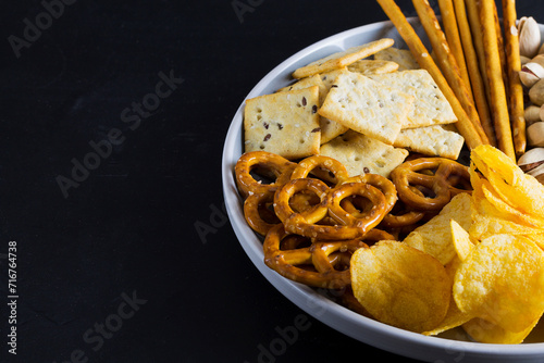 Crackers, potato chips and mini pretzels in the grey plat. Assortment of crispy appetizers. Salty snacks on a black background. Party mix. copy space