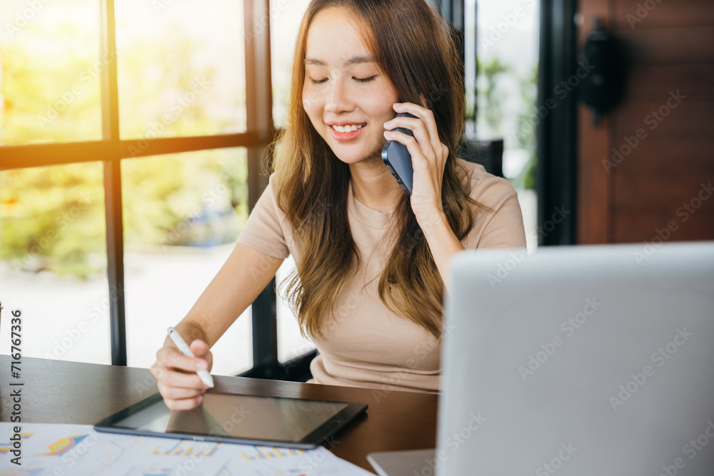 Smiling female working laptop computer and take note on digital tablet at home office, Happy freelance Asian woman sitting at cafe call mobile phone talking and asking about something, financial