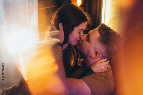 A couple of a guy and a girl gently hug and kiss in a room lit with colored warm light among yellow highlights.