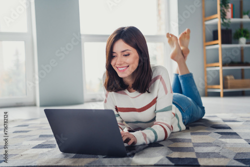 Full size photo of cute cheerful girl laying carpet floor use wireless laptop typing email living room house inside