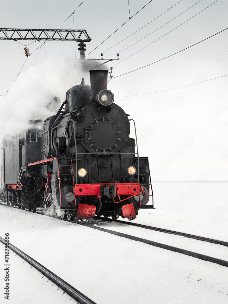 Fototapeta premium Old steam engine locomotive driving by in a snowy field. Black vintage locomotive pulling a train on a winter day. 