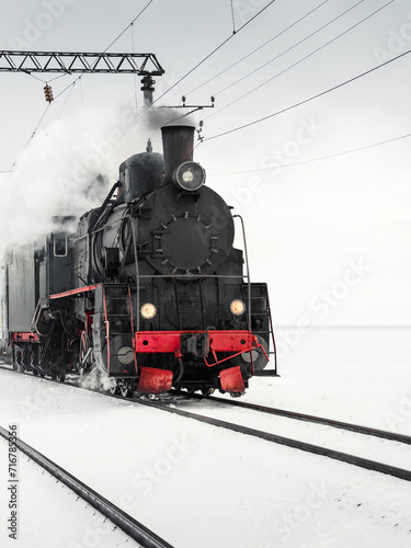 Old steam engine locomotive driving by in a snowy field. Black vintage locomotive pulling a train on a winter day. 