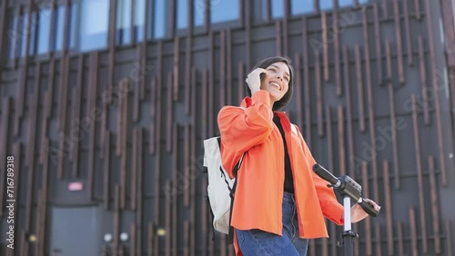 Lady expressing positive emotions during energetic smartphone call on contenporary electric scooter outdoors. Emotional caucasian woman showing hello gesture to person who far away. photo