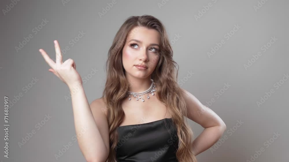A beautiful young girl with makeup, brown hair in a black spaghetti strap dress mentally counts something on her fingers while posing in the studio on a gray background. A beautiful young girl with makeup, brown hair in a black spaghetti strap dress mentally counts something on her fingers while posing in the studio on a gray background.