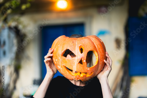 Caucasian Adult Woman Looking Trough a Handmade Carved Jack 'O' Lantern for Upcoming Halloween Night Outdoors in Front of her Countryside Blue Windows and Doors House