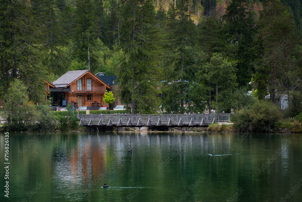 Ein von Bäumen umgebenes Holzhaus an einem Seeufer vor einem Wald, mit einem Metallsteg über den See