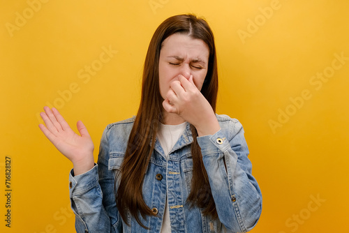 Portrait of sad young woman standing pinching her nose with fingers to hold breath, disgusted by stinky intolerable smell, wearing denim jacket, isolated on yellow studio background. Bad smell concept