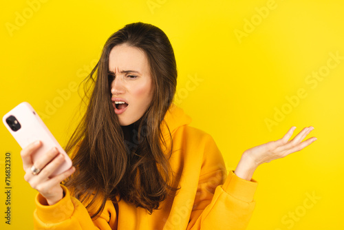 Mad and shock young brunette beautiful woman using mobile phone isolated on yellow background. Yelling unhappy woman hold mobile phone. Disappointed sad upset lady horrified impressed news. WTF. Oh no photo