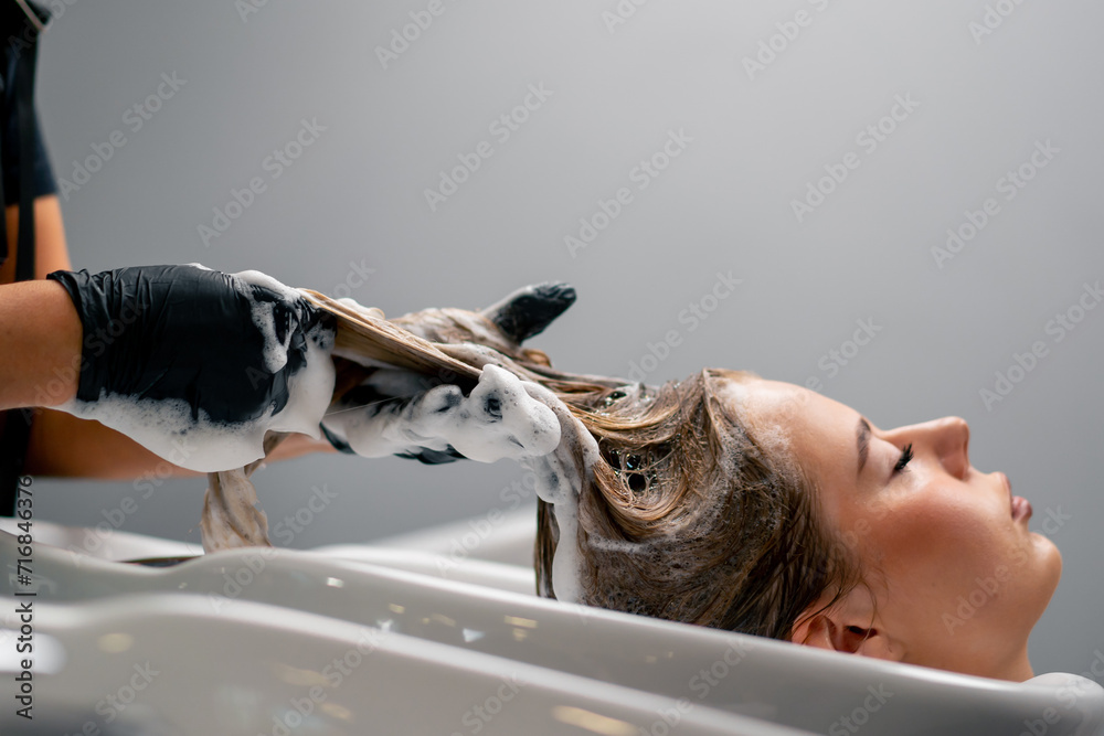 a hairdresser washing off dye from a client's hair during a care procedure in a beauty salon - obrazy, fototapety, plakaty 