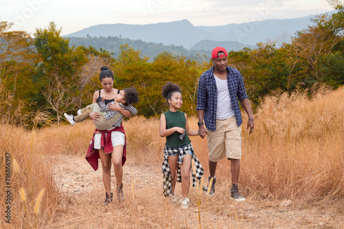 Happy African American family with adorable children weekend vacation hiking outdoors in countryside, father and mother carry children walking trail in forest park with fun, bonding relationship